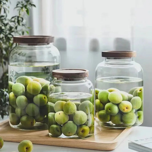 Three glass jars with wooden lids filled with green apples on a wooden tray.