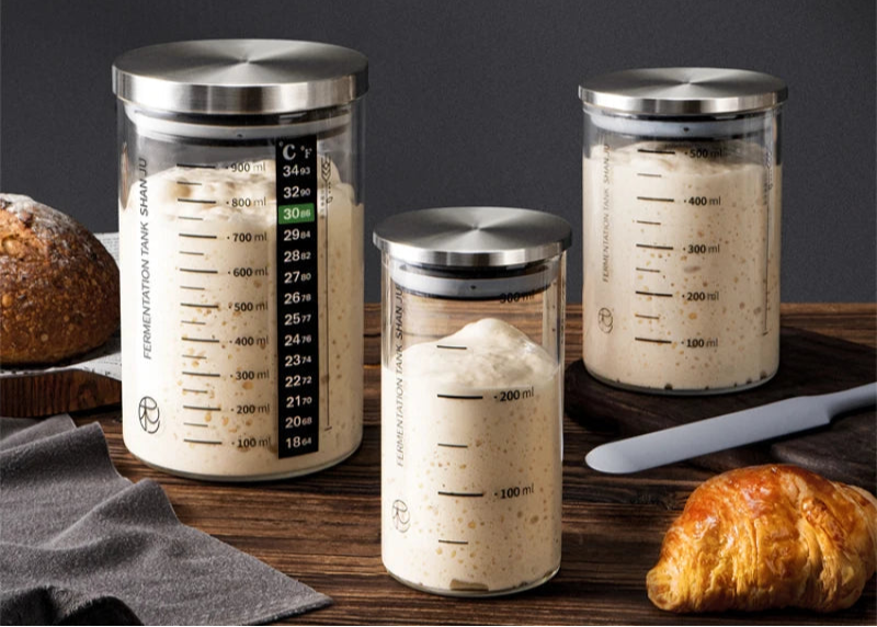 Three glass containers with metal lids on a wooden surface with bread and a knife.
