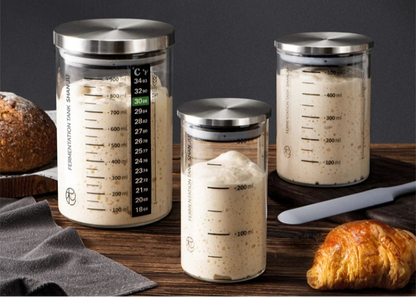Three glass containers with metal lids on a wooden surface with bread and a knife.