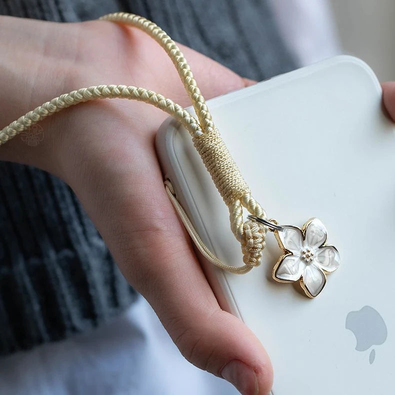 Hand holding a white phone case with a gold cord and flower charm, blurred background