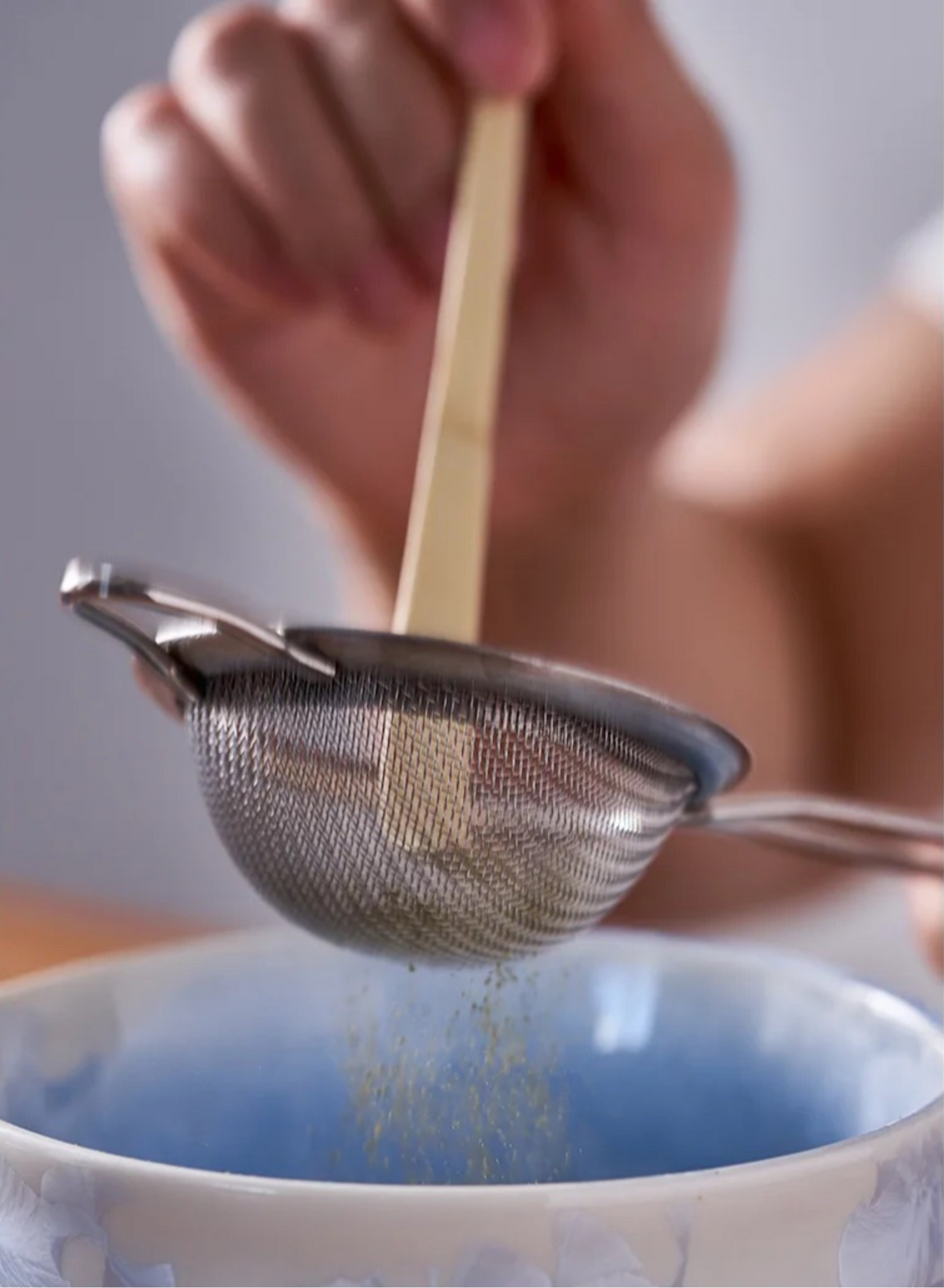 Hand using a small mesh strainer over a bowl with a wooden spoon