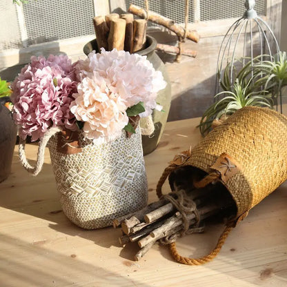 Decorative setting with a textured vase holding flowers, a woven basket, and sticks on a wooden surface.