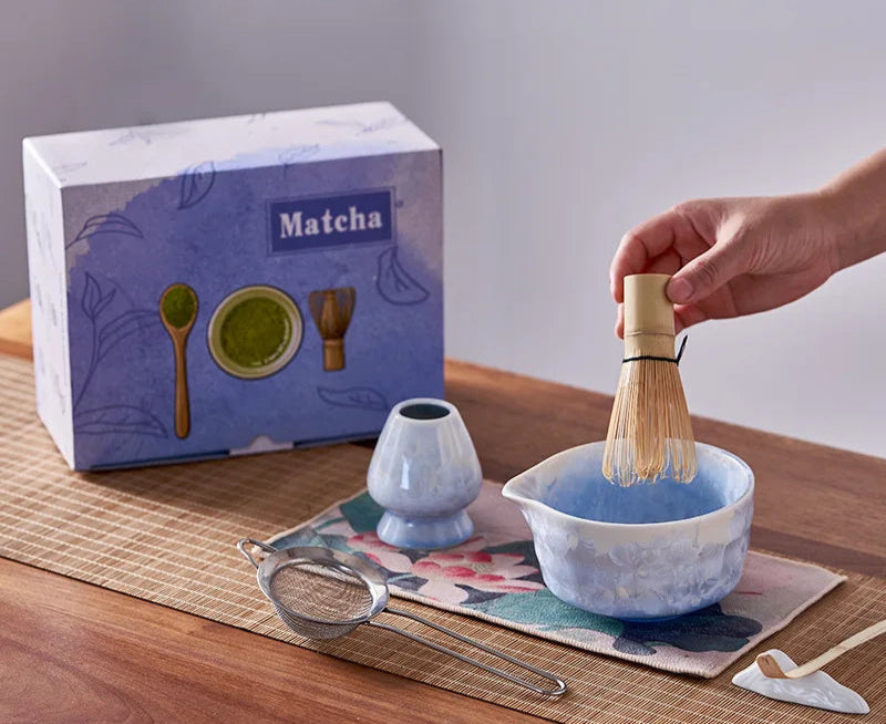Matcha tea set with a hand holding a whisk, a box labeled 'Matcha', and a bowl on a wooden surface.
