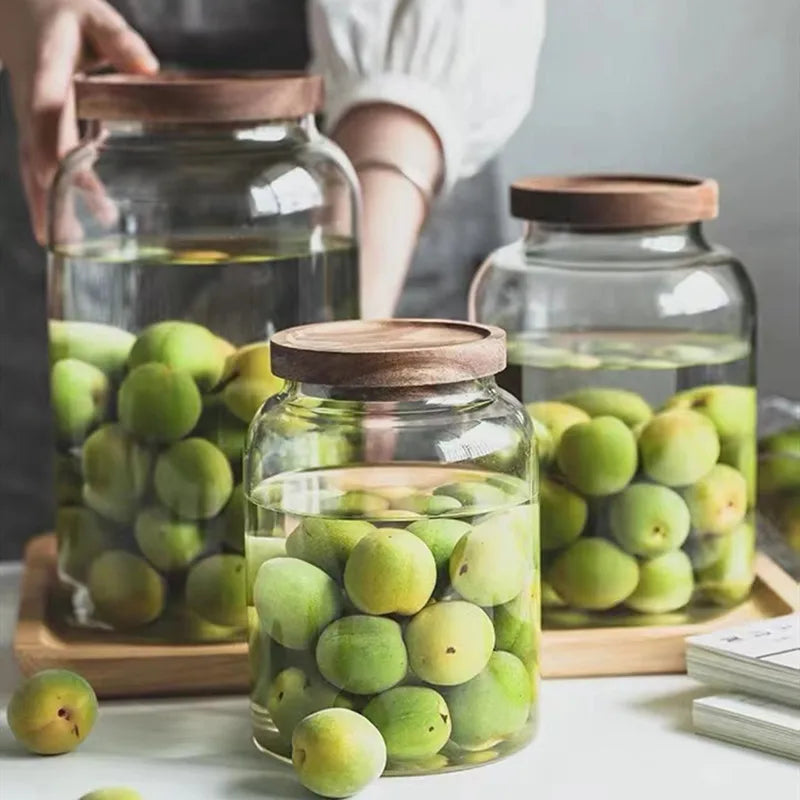 Three glass jars with wooden lids filled with green plums on a wooden tray.