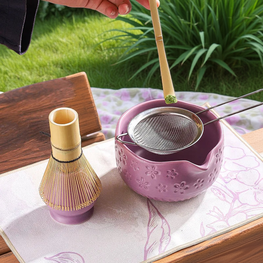 Person making matcha tea spooning matcha into a pink bowl on a wooden table with plants in the background.