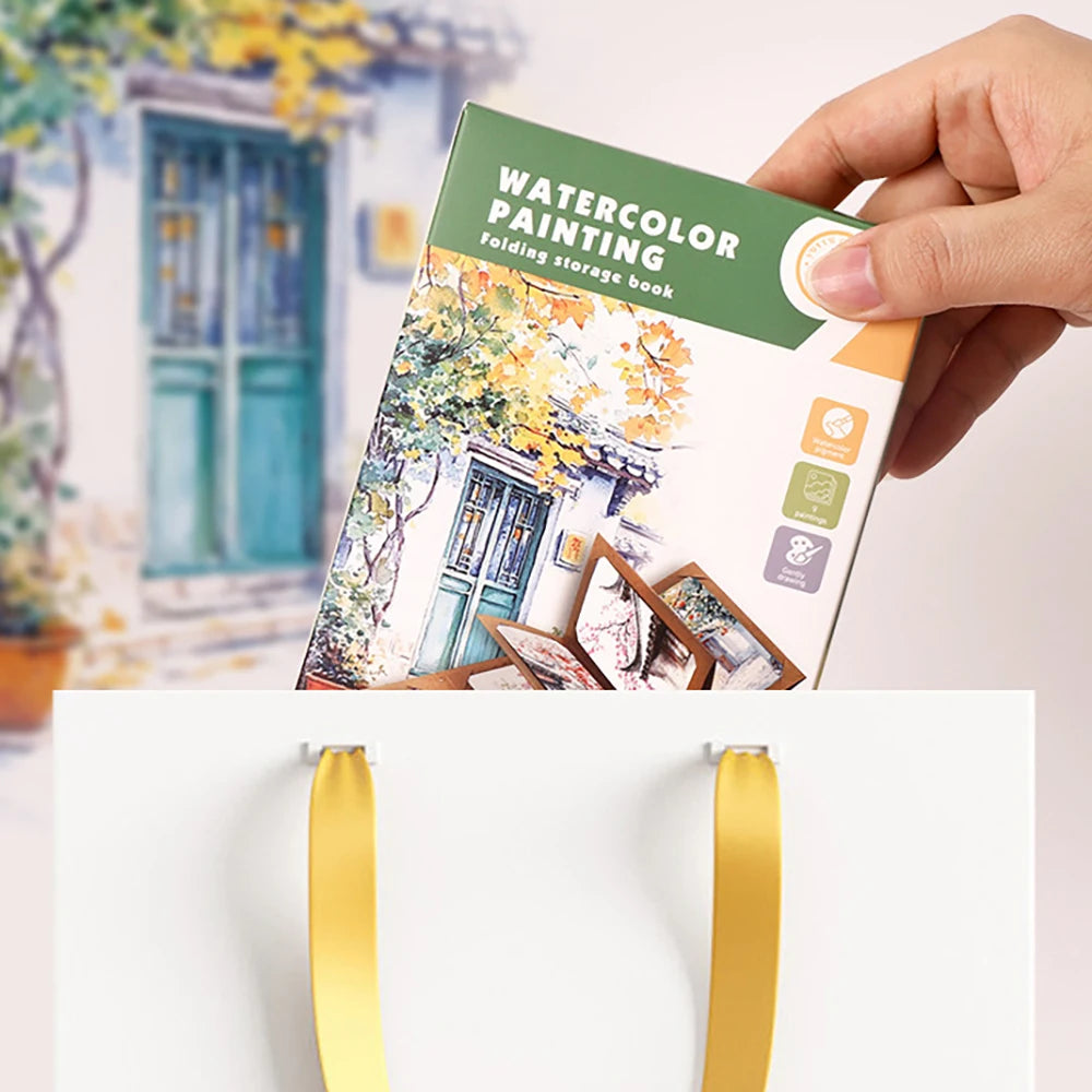 Hand holding a watercolor painting book with a scenic cover, placed on a white surface with yellow ribbons.