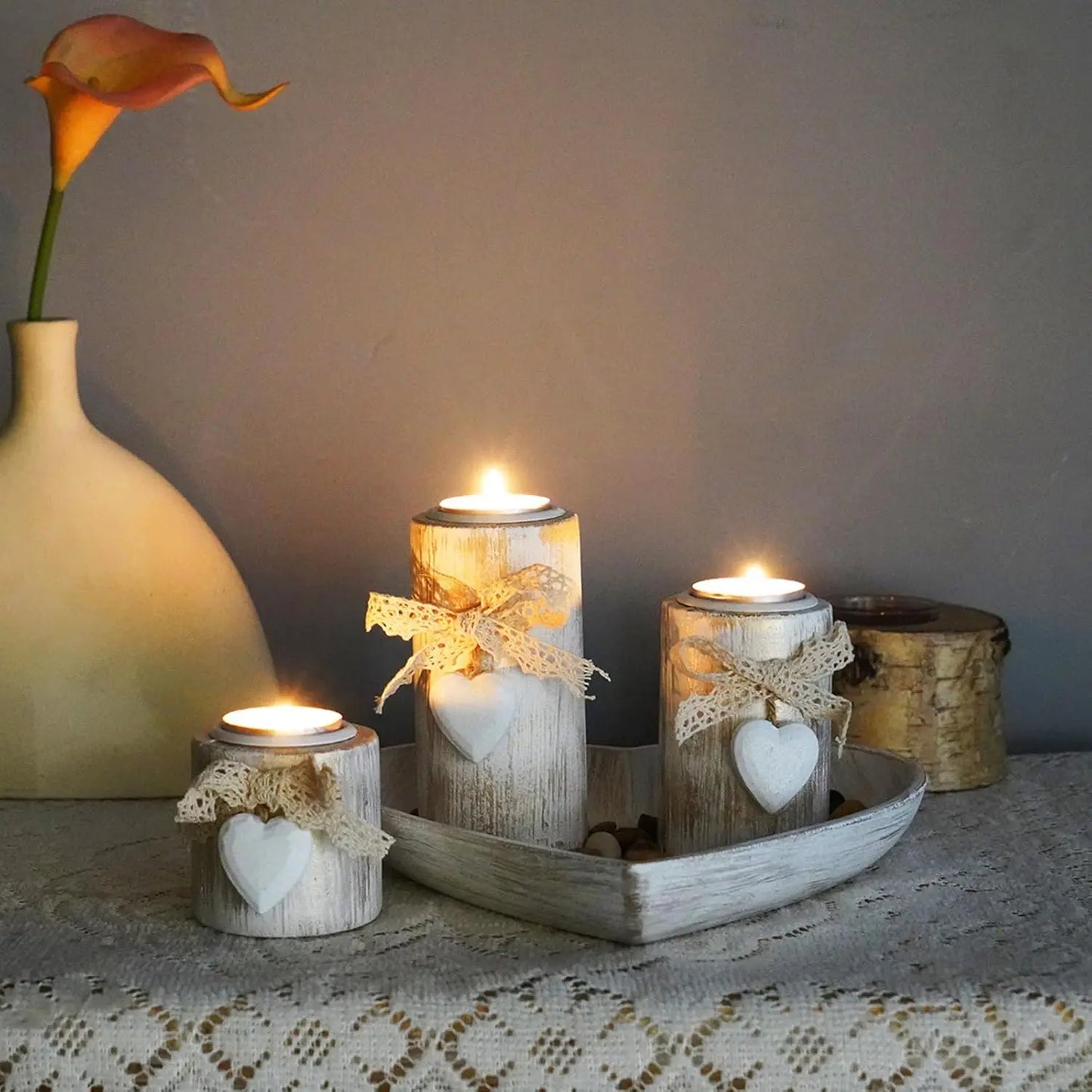 Decorative candles with heart designs on a lace tablecloth, with a vase and flower in the background.
