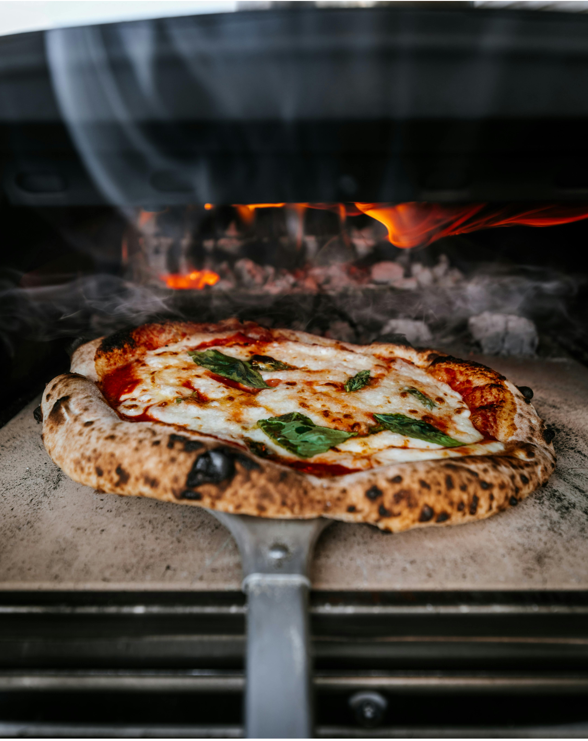Pizza being cooked in a wood-fired oven with flames visible.