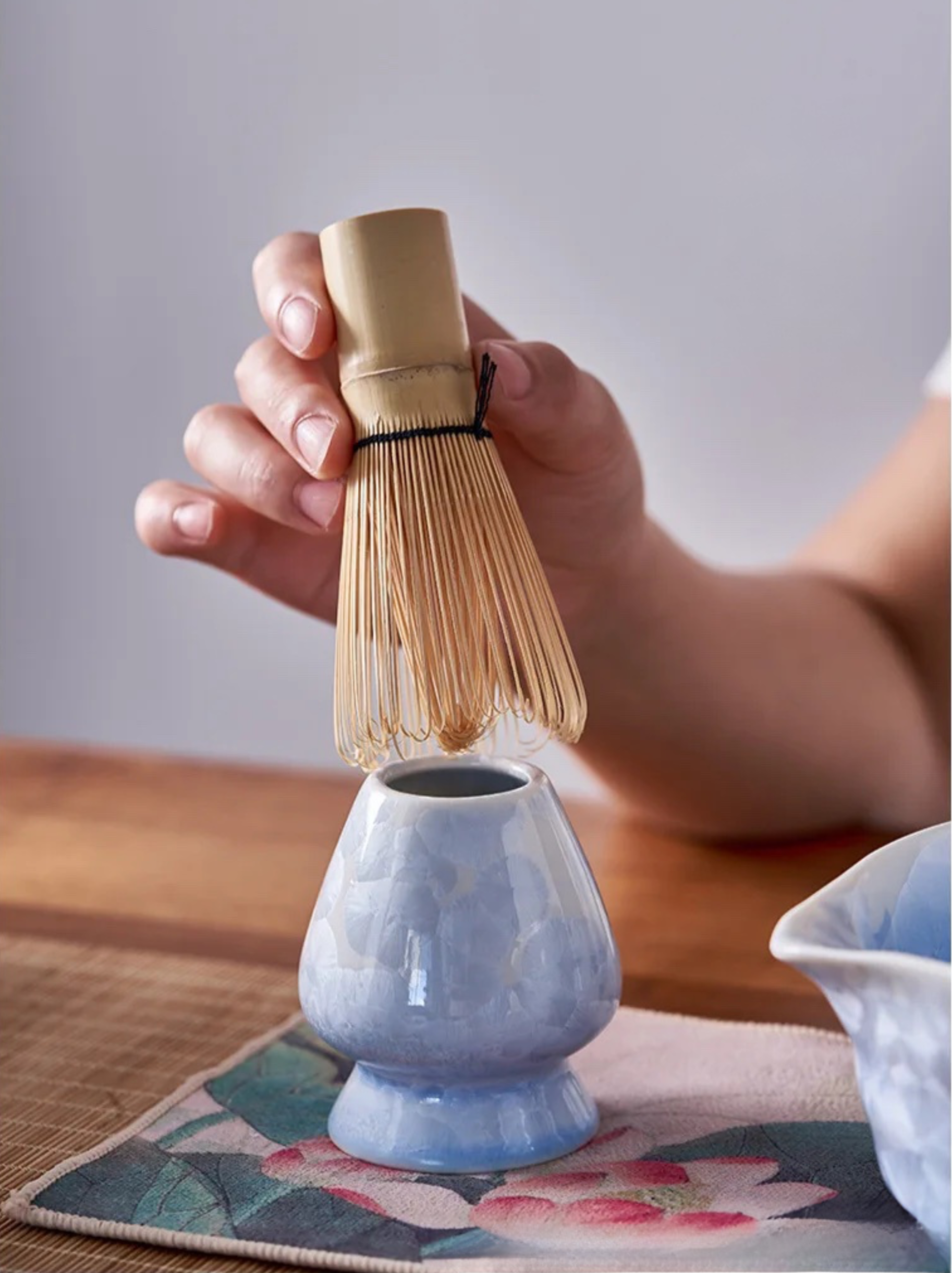Hand holding a wooden whisk above a small blue ceramic cup on a wooden table.