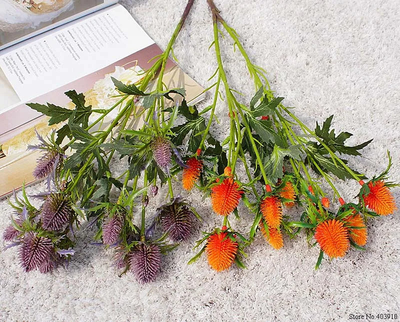 Artificial flowers with green leaves and orange and purple buds on a textured surface.