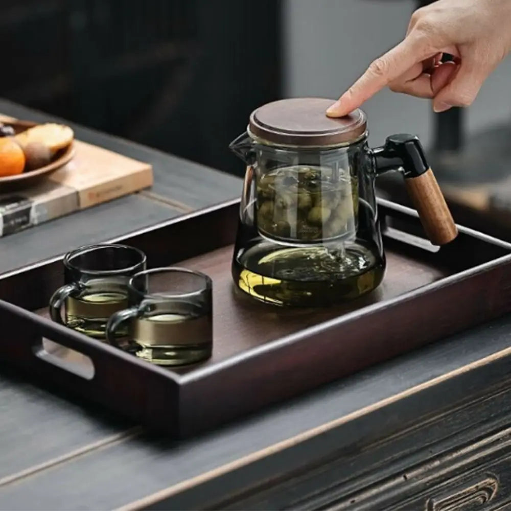 Tea set with glass teapot and cups on a wooden tray, hand pointing at the teapot.