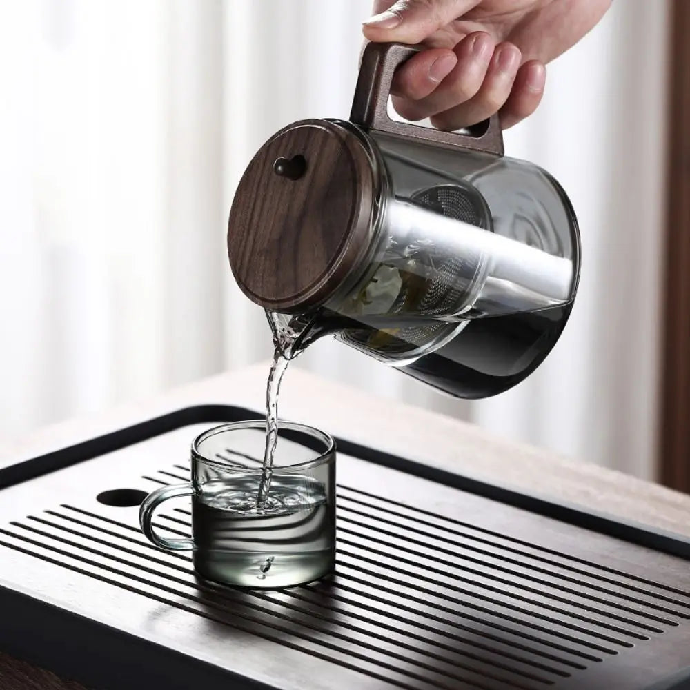 Glass teapot with wooden lid being poured into a glass on a tray.