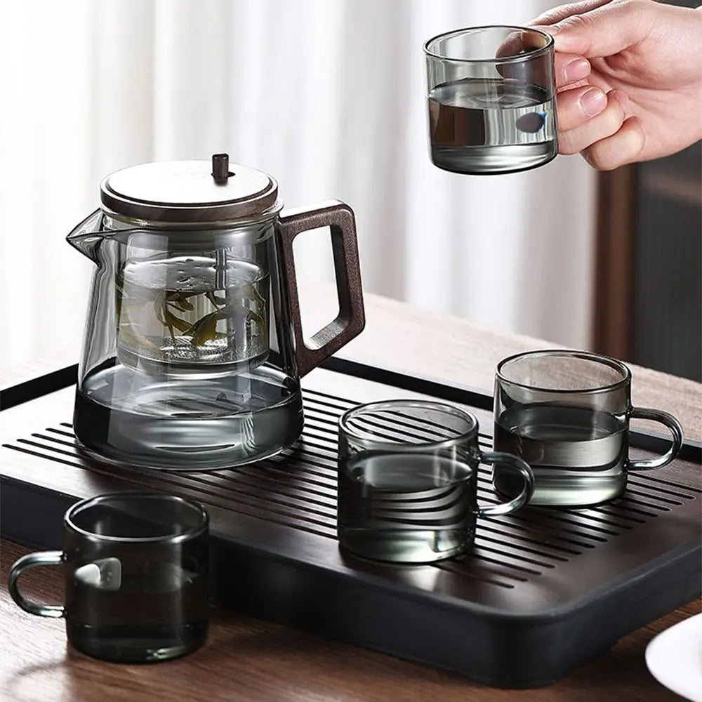 Glass teapot and cups on a tray with a hand holding a cup of water.