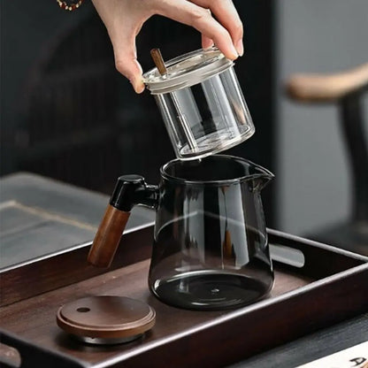 Glass teapot with wooden handle on a wooden tray, hand holding a clear glass container.