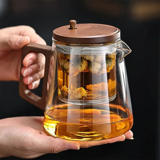Glass teapot with wooden lid and handle, filled with tea and flowers, held by a hand against a dark background.