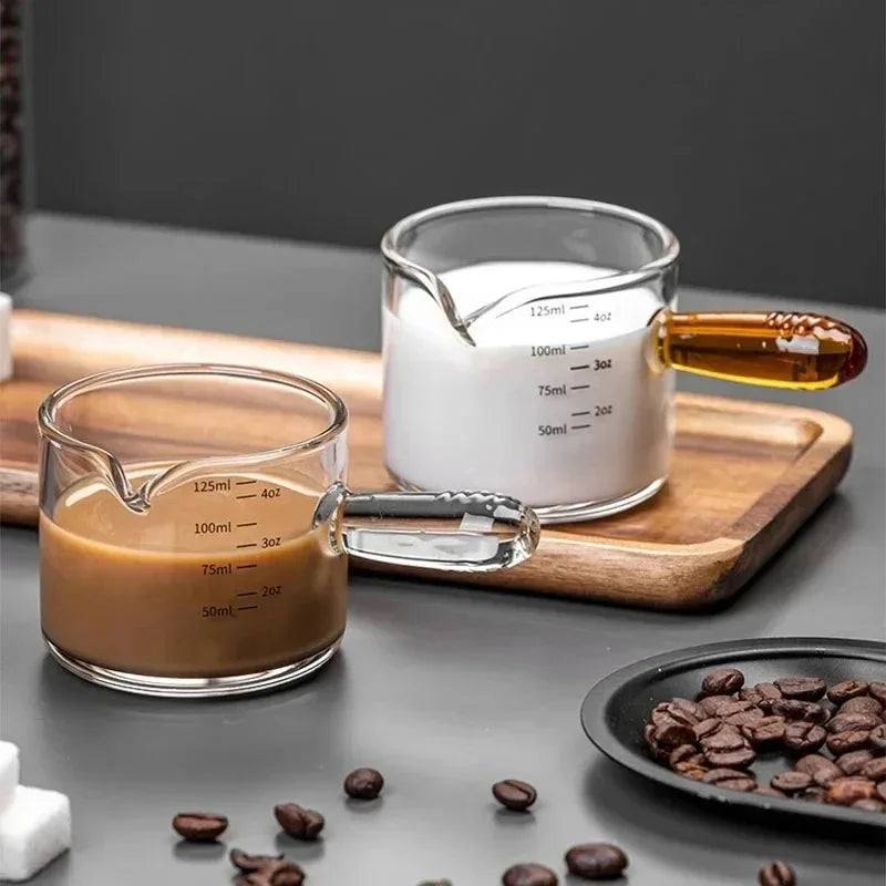 Two glass measuring cups with coffee and milk, surrounded by coffee beans on a dark surface.