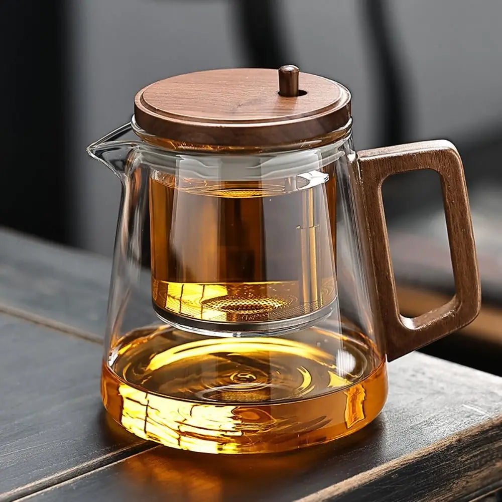 Glass teapot with wooden lid and handle filled with tea on a wooden surface.