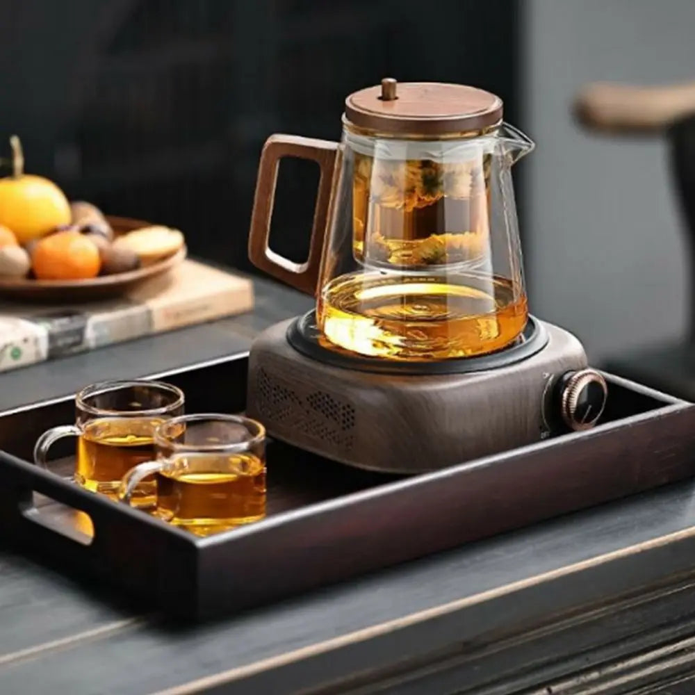 Glass teapot with wooden handle on a hot plate, accompanied by two cups of tea on a tray.