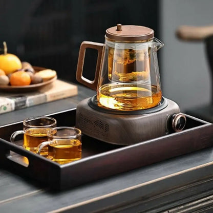 Glass teapot with wooden handle on a hot plate, accompanied by two cups of tea on a tray.