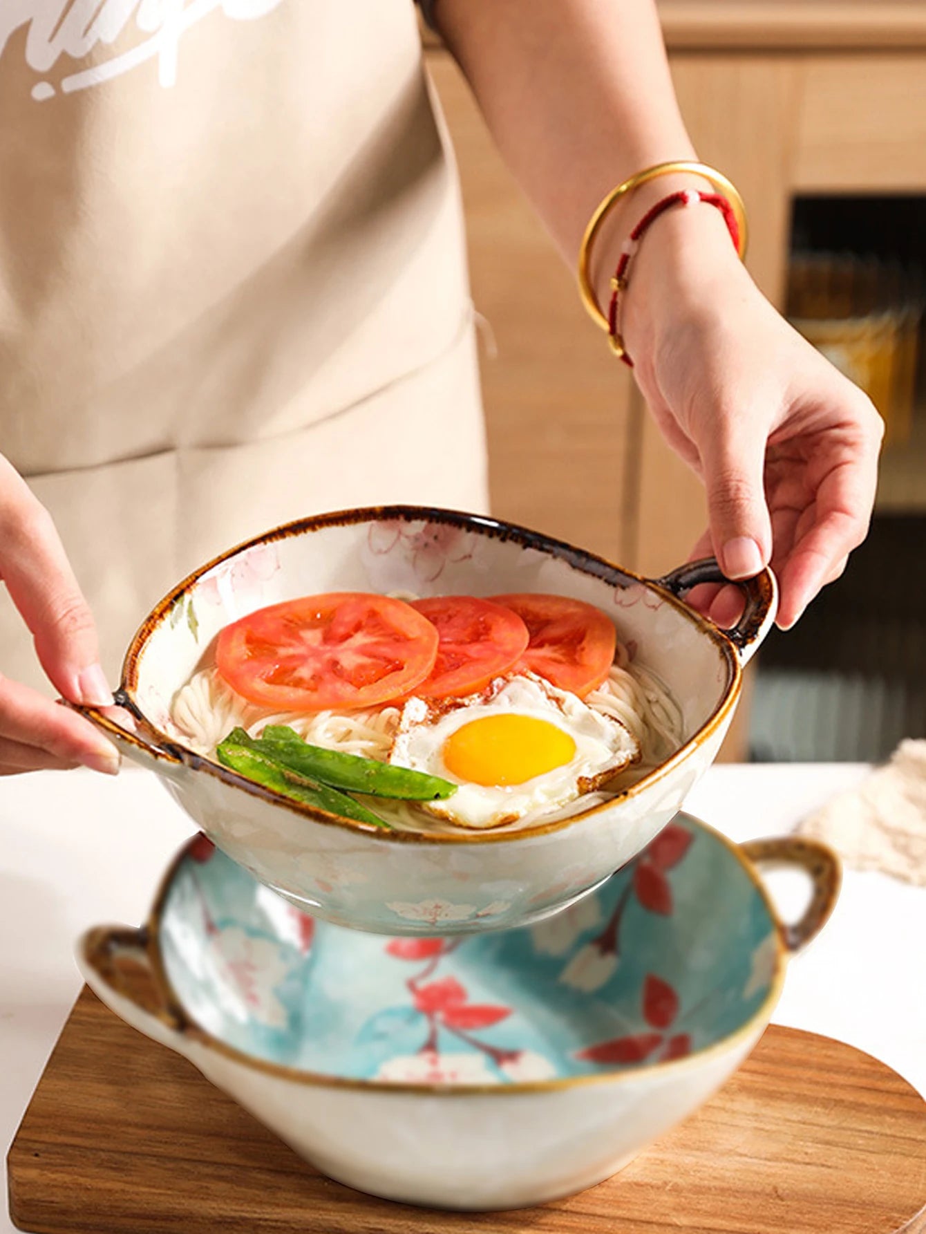 Person holding a bowl of ramen with tomatoes and an egg on a wooden table.