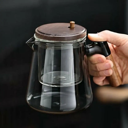 Glass teapot with wooden lid and handle held by a hand against a dark background