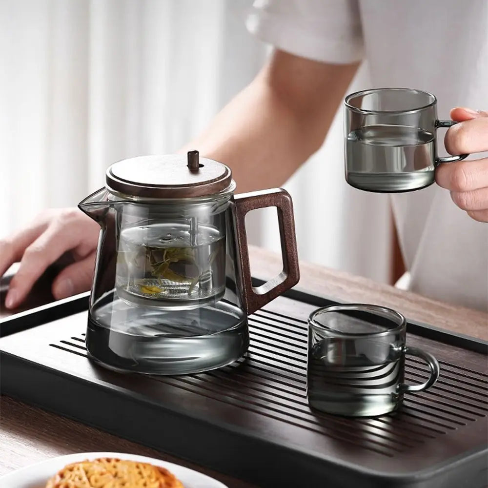Glass teapot and cups on a wooden tray with a person holding a cup.
