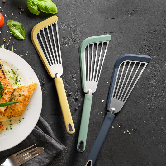 Three kitchen spatulas with different colored handles on a dark surface with a plate of food.