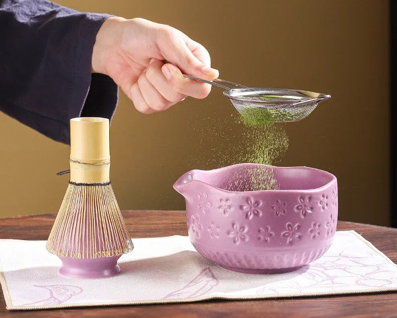 Person sifting green tea powder into a purple bowl with a bamboo whisk on a wooden surface.
