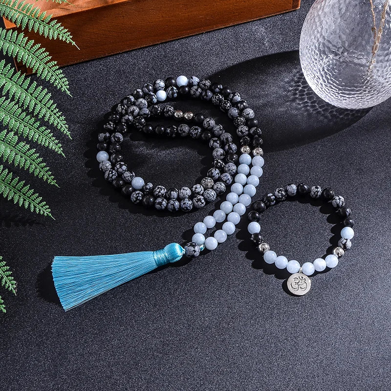 Mala beads with a blue tassel and OM charm on a dark surface with greenery.