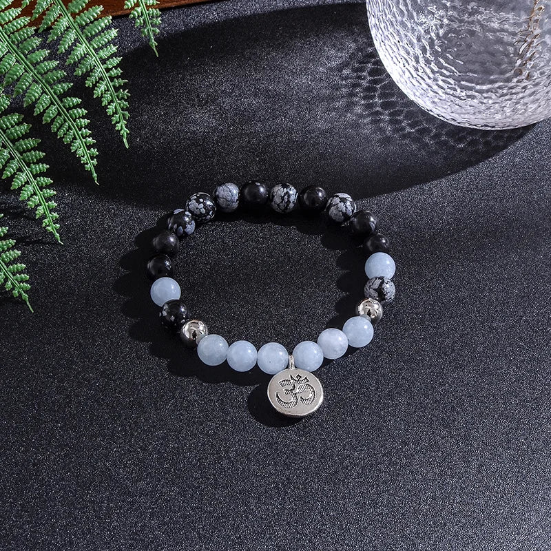 Beaded bracelet with a centralOm symbol charm on a dark surface with ferns and a glass.