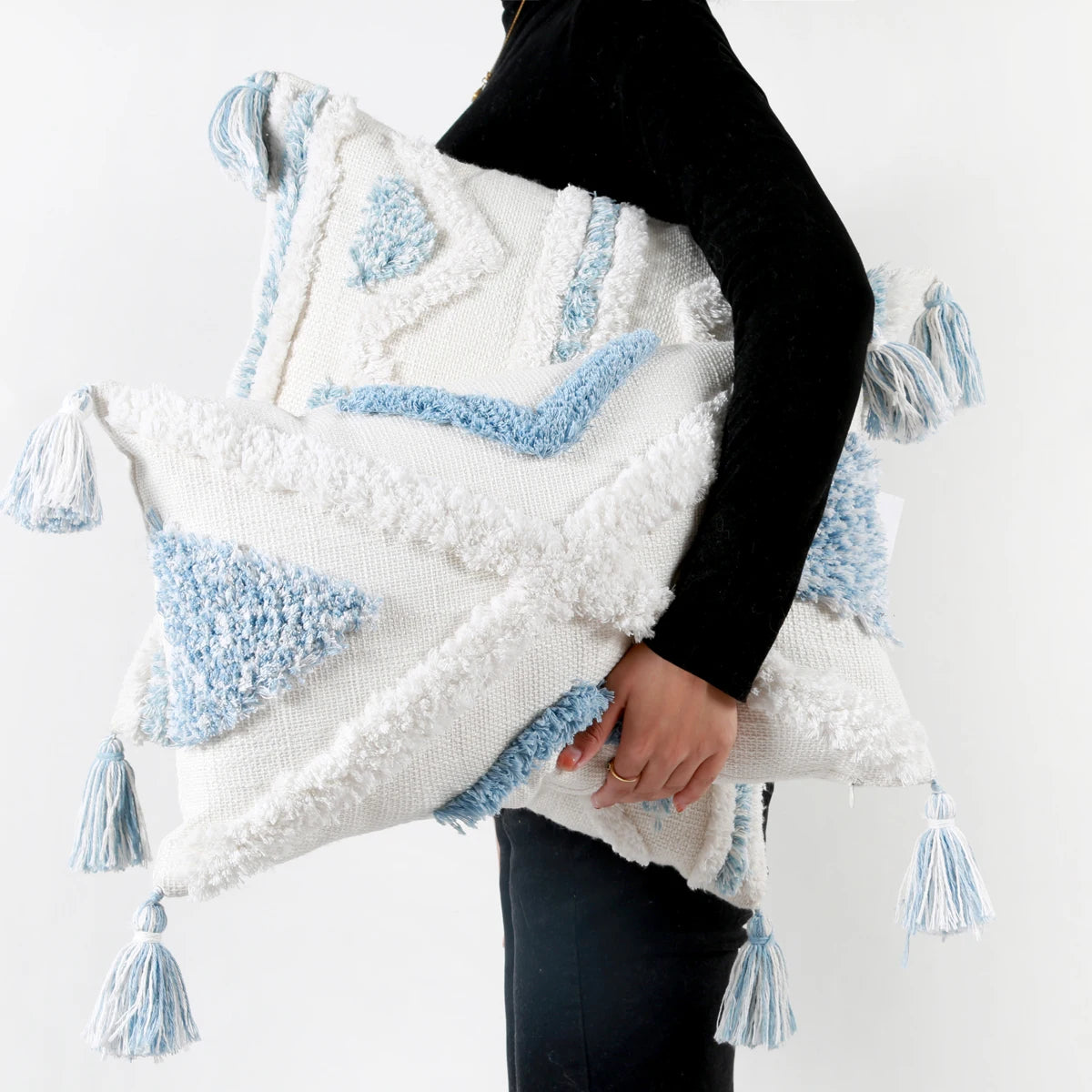 Person holding a decorative pillow with white and light blue patterns and tassels on a plain background