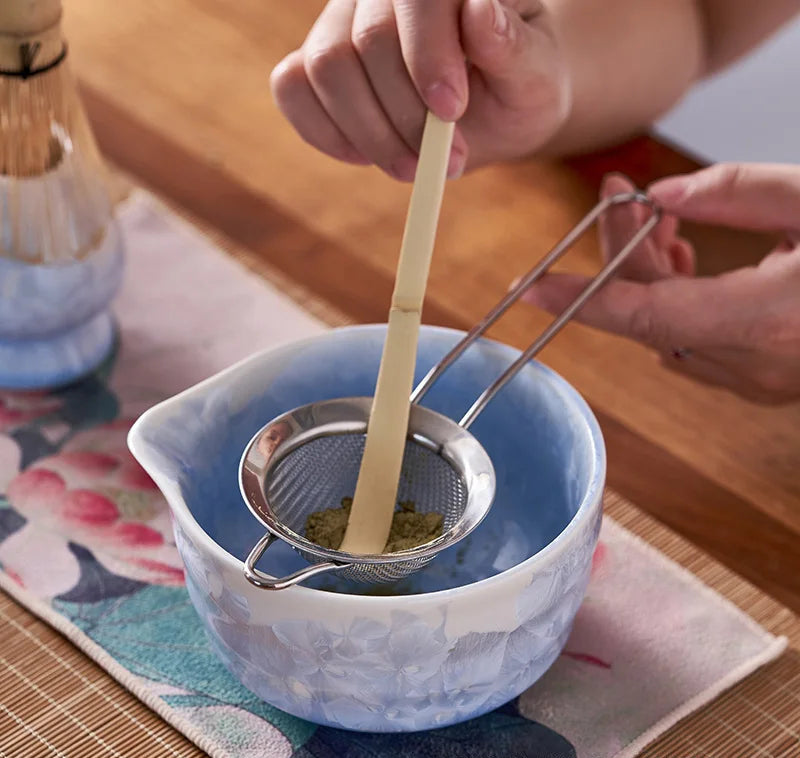 Person using a tea strainer in a blue ceramic bowl on a wooden table.