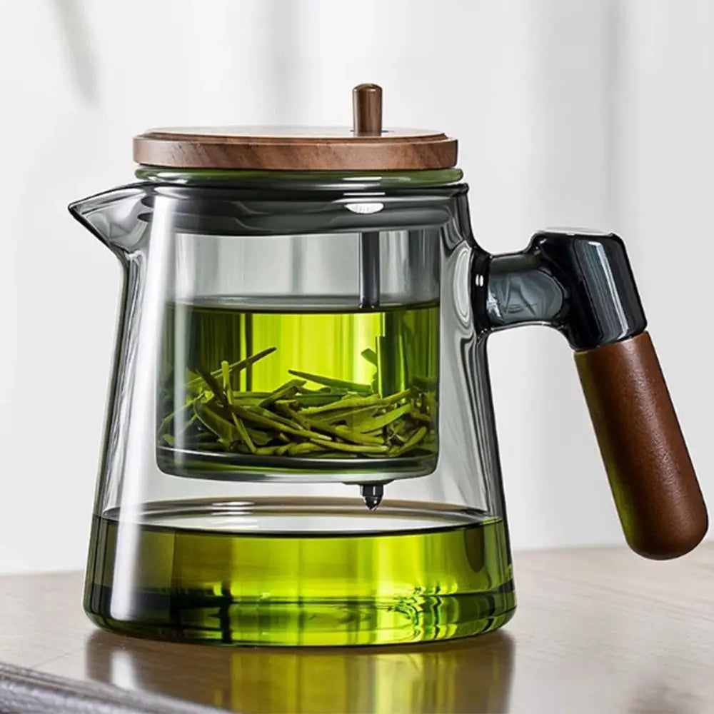 Clear glass teapot with wooden lid and handle filled with green tea leaves on a light background