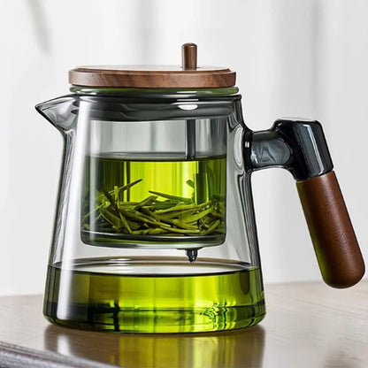 Clear glass teapot with wooden lid and handle filled with green tea leaves on a light background