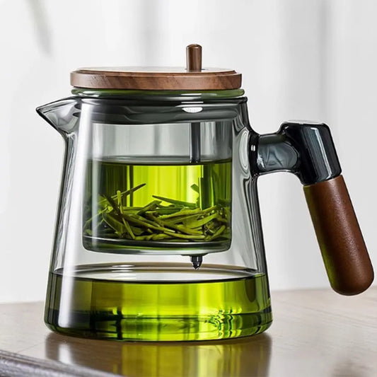 Clear glass teapot with wooden lid and handle filled with green tea leaves on a light background