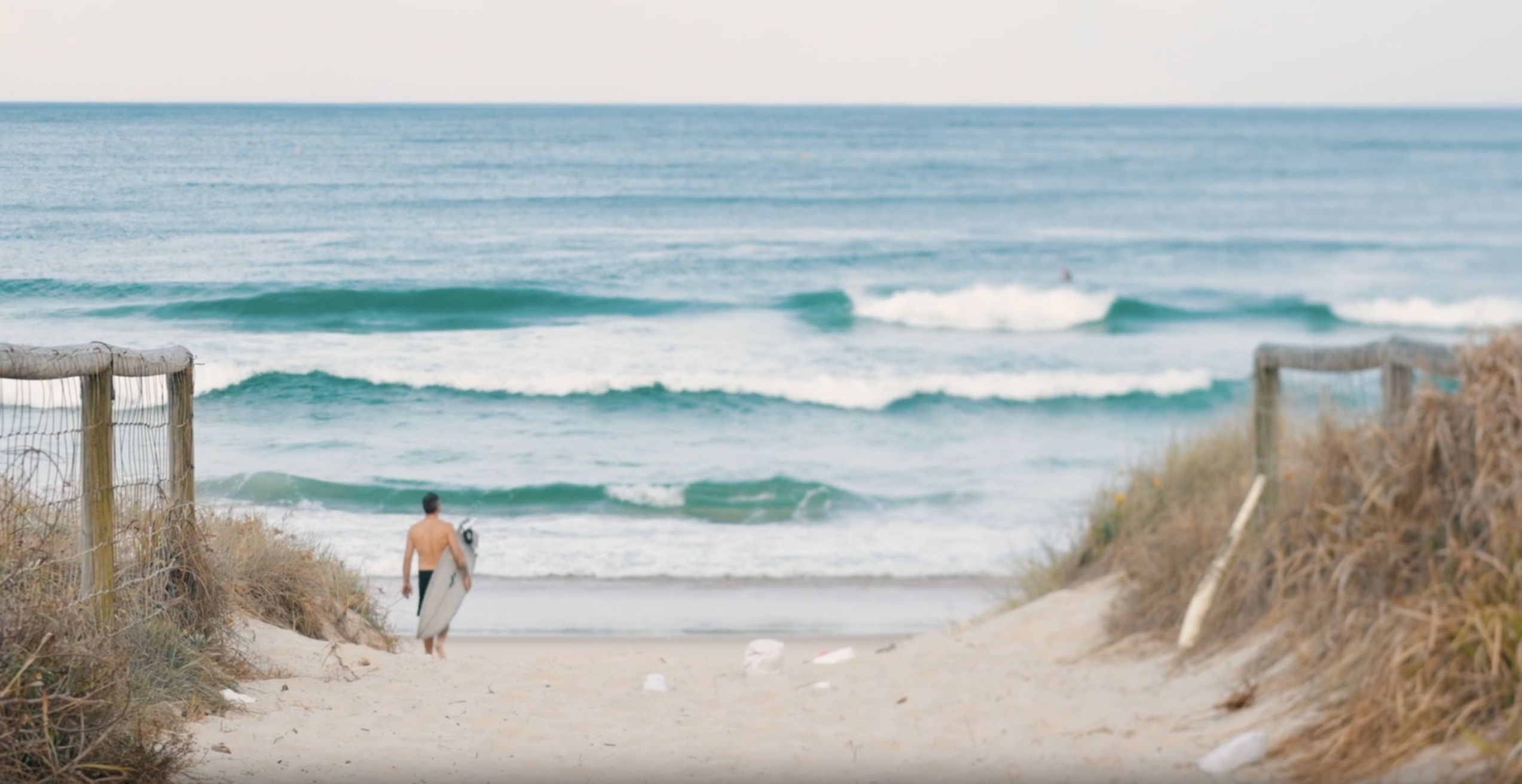 Load video: Person walking along a sandy beach path toward the ocean, carrying a surfboard with gentle waves in the background.