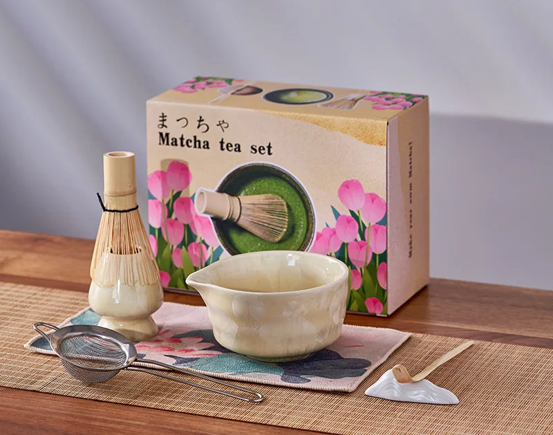 Matcha tea set with bowl, whisk, and other tools on a wooden surface with a floral box in the background.