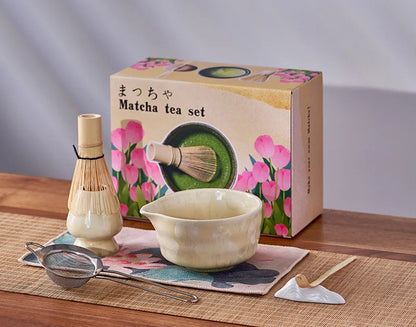 Matcha tea set with bowl, whisk, and other tools on a wooden surface with a floral box in the background.