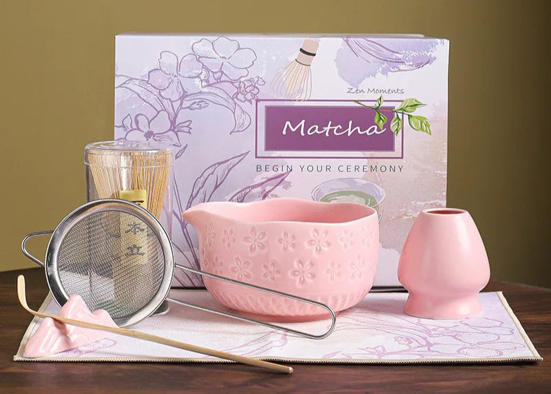 Matcha tea set with pink bowl, small cup, and strainer in front of a Matcha-themed book on a wooden surface.