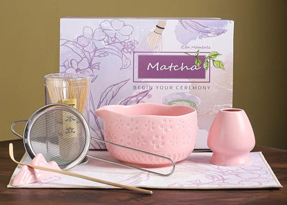 Matcha tea set with pink bowl, small cup, and strainer in front of a Matcha-themed book on a wooden surface.