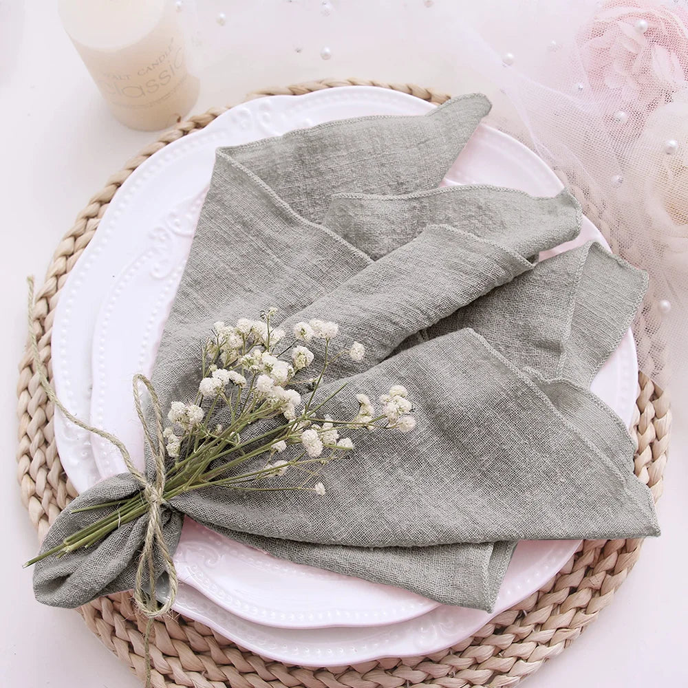 Folded gray napkin with a small bouquet of white flowers on a white plate with a pink cloth, all placed on a woven mat.