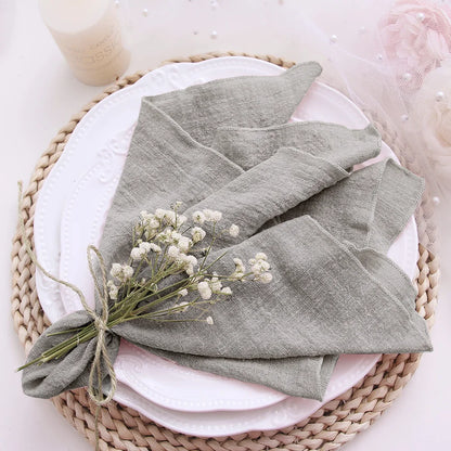 Folded gray napkin with a small bouquet of white flowers on a white plate with a pink cloth, all placed on a woven mat.